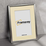 Rectangle silver poster frame with ivory mount on a floor leaning with the wall with a Framorey logo placed at the frame's center.