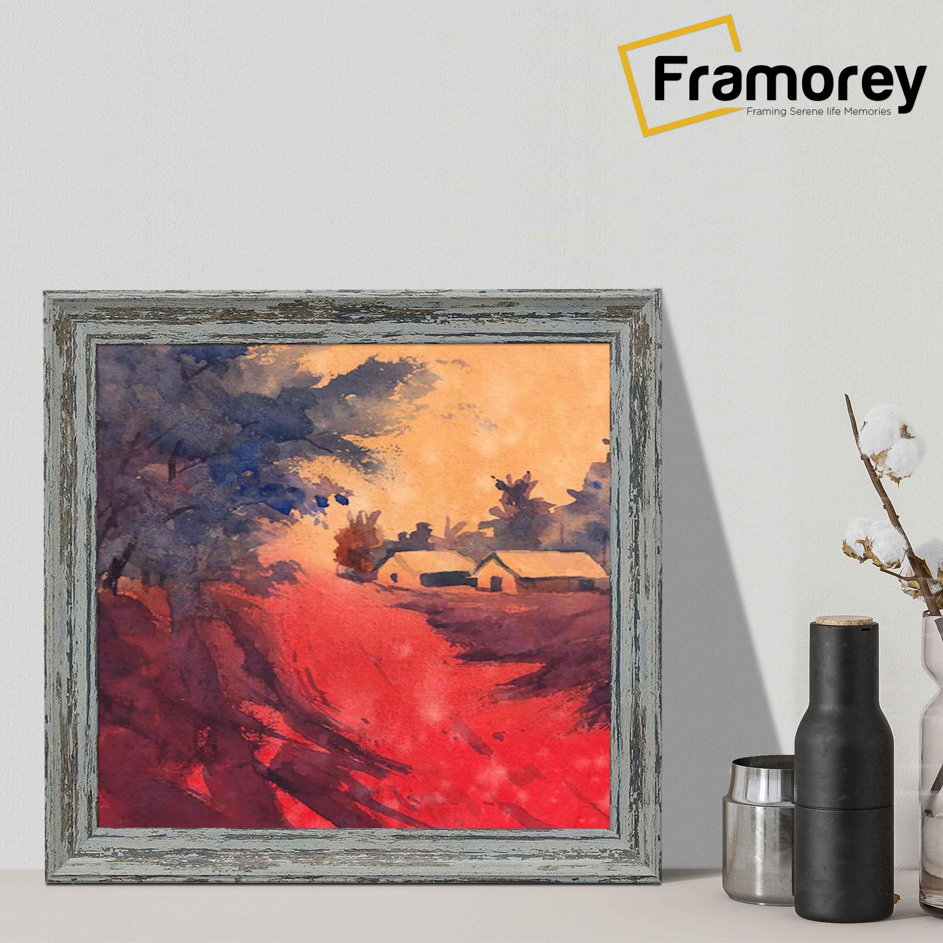 Framed watercolor painting depicting a rural landscape with houses, trees, and a vivid red pathway under a warm sunset sky. The frame has a rustic, weathered wood finish. The Framorey logo is positioned in the top right corner, and decorative vases are placed beside the frame on a neutral-colored surface.