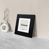 Side view of a Square Black Photo Print Frame With White Mount with a Framorey logo and the frame is leaning with the wall on a wooden table.