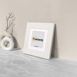 Side view of a Square White Photo Print Frame with White Mount with a Framorey logo place in the frame and the frame is leaning with the wall.